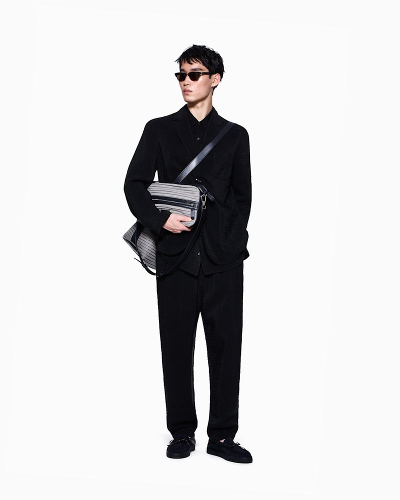 Man wearing a black suit and sunglasses carries a striped messenger bag against a plain white studio background.