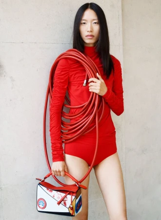 An Asian woman in a red bodysuit poses in front of a concrete wall, holding a hose and a patterned handbag in a fashion shot.
