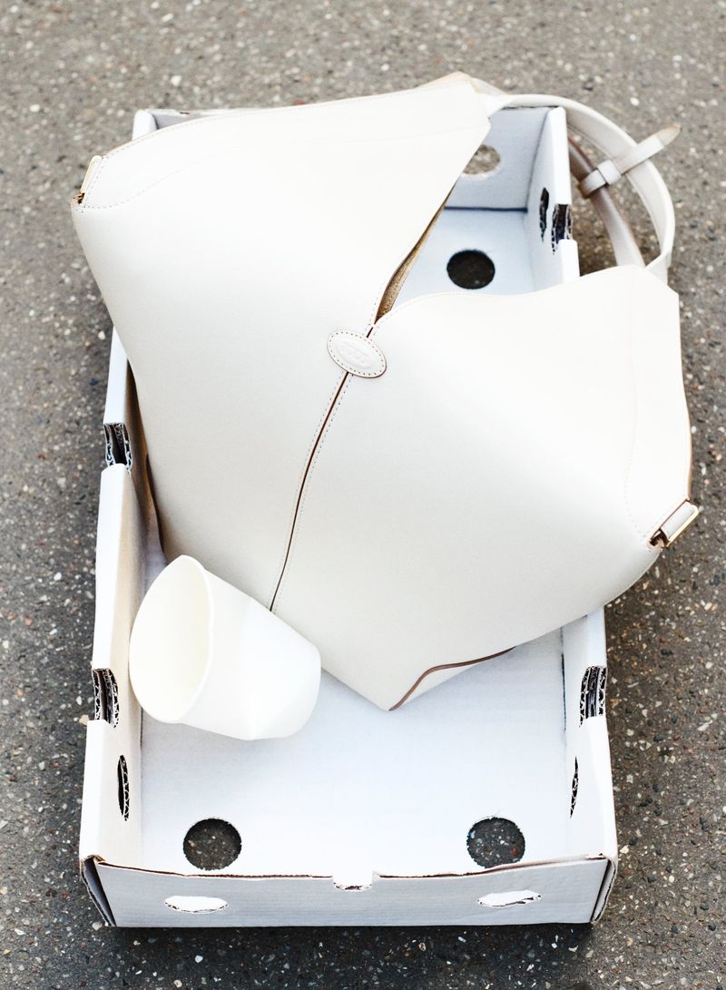 An open white cardboard box contains a white cup and a white purse, captured in an overhead shot outdoors on a neutral surface.