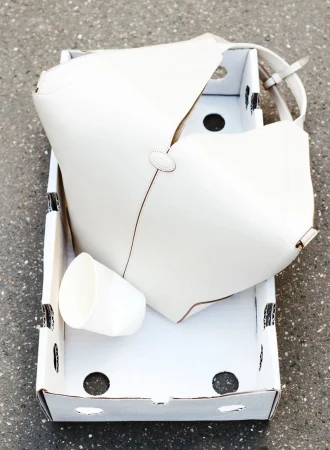 An open white cardboard box contains a white cup and a white purse, captured in an overhead shot outdoors on a neutral surface.