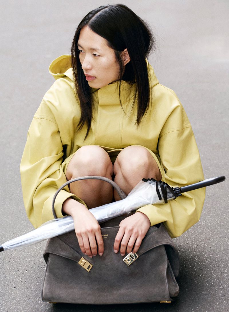 Asian woman crouches on asphalt with gray purse and umbrella. She wears yellow raincoat. The image shows a fashion forward aesthetic.