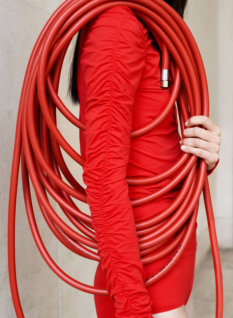 Woman in red dress poses holding a large, coiled red garden hose in a portrait-style shot in front of a neutral background.