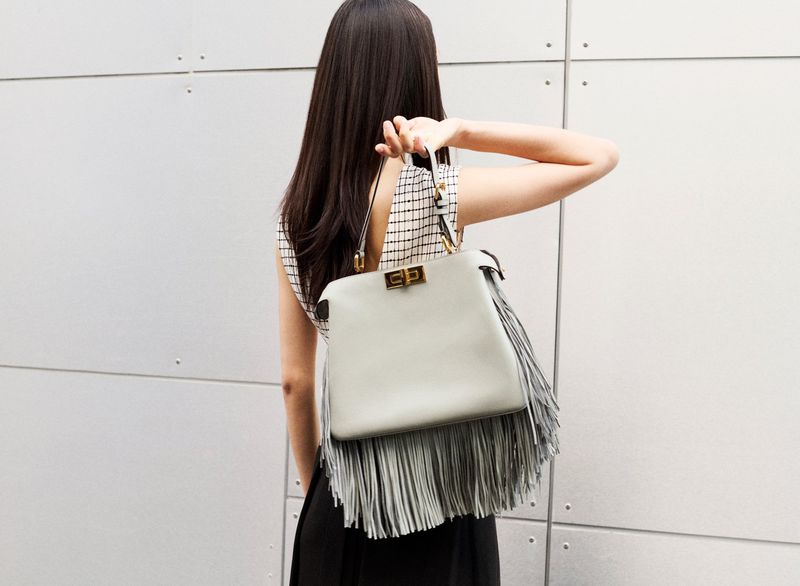 Woman carries a light grey fringe purse. The bag features a rectangular shape and is carried on her shoulder indoors against a grey wall.