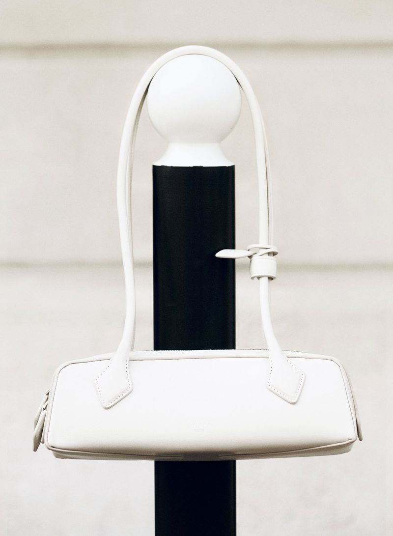 Ivory-colored leather baguette bag with thin handle hanging on a black and white post outdoors in neutral lighting.