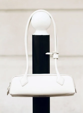 Ivory-colored leather baguette bag with thin handle hanging on a black and white post outdoors in neutral lighting.
