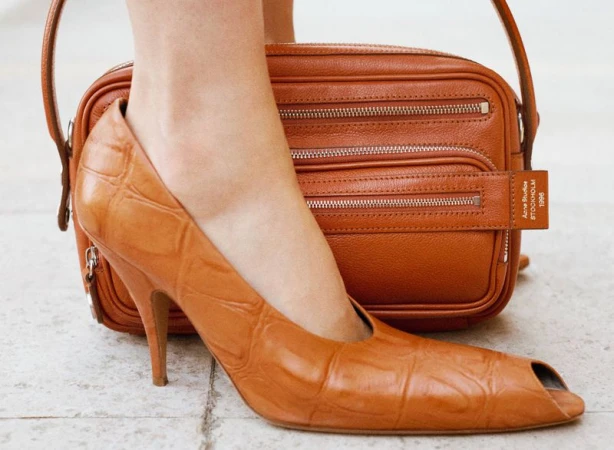 Close-up of a caramel-colored open-toed high heel with a matching leather purse in soft studio lighting on a marble floor.
