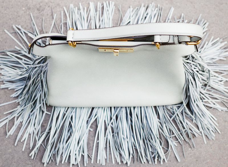 White leather purse with fringe and a gold clasp rests on a gray surface, showcasing a stylish accessory.
