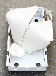 Cream leather tote bag and a white paper cup arranged inside a white cardboard crate on dark asphalt ground.