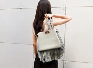 Model seen from behind holding a pale grey fringed handbag against a tiled wall