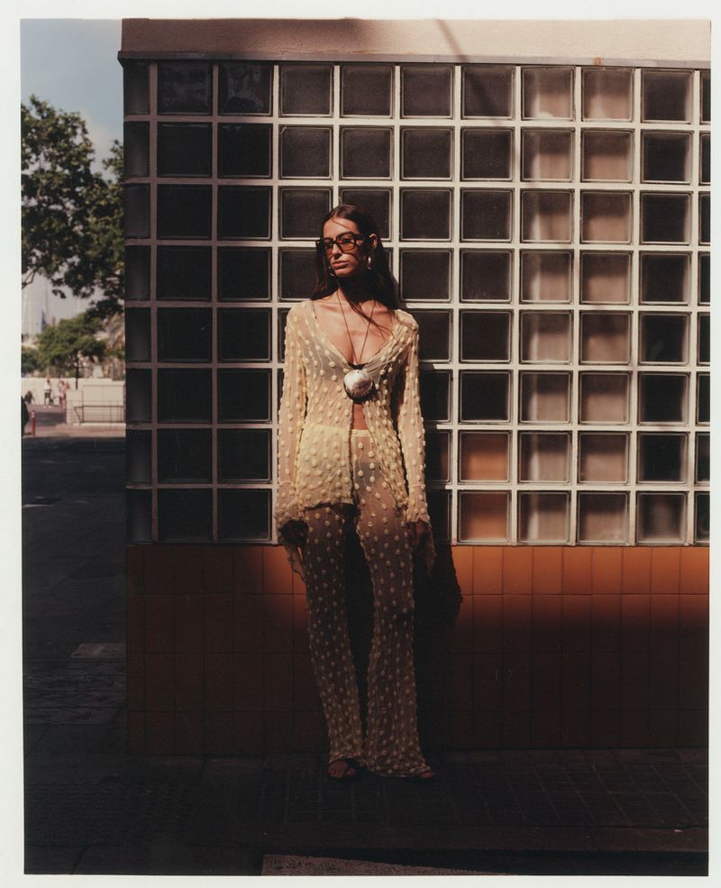 Woman in sheer floral bell-bottom pantsuit and sunglasses posing in front of a glass block wall with a white frame around it.