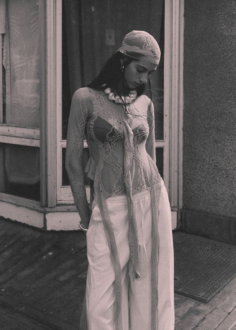 Model wearing a sheer lace long-sleeve top, matching headscarf, and wide-leg trousers standing outside in black and white.
