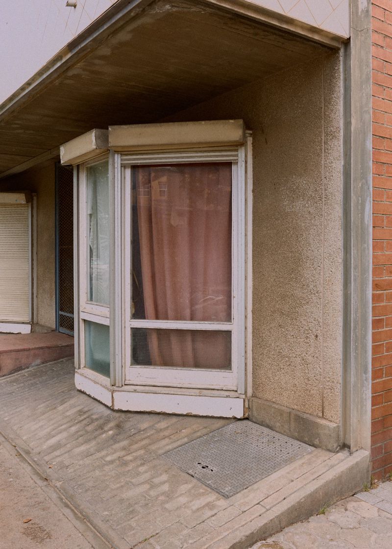 Exterior view of a building featuring a distinctive white-framed window with drawn curtains, showcasing architecture with concrete and brick elements.