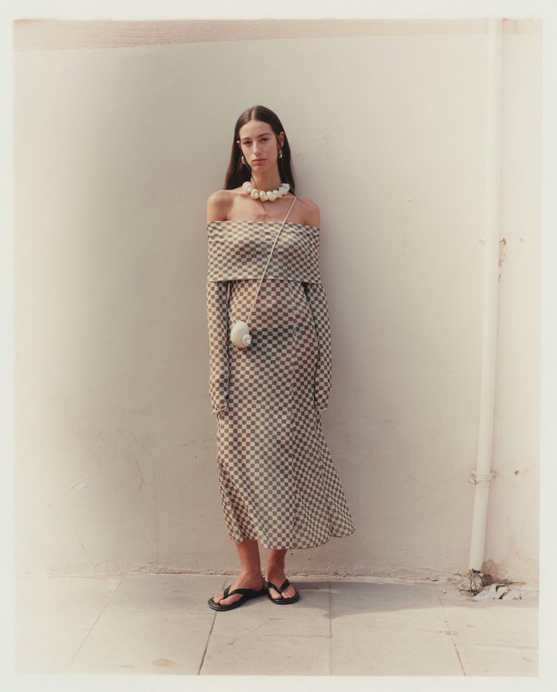 Elegant adult woman poses against a wall in a checkered off-the-shoulder dress, pearl necklace, and sandals for fashion shoot.