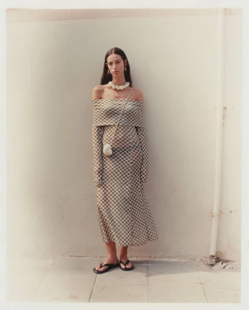 Elegant adult woman poses against a wall in a checkered off-the-shoulder dress, pearl necklace, and sandals for fashion shoot.