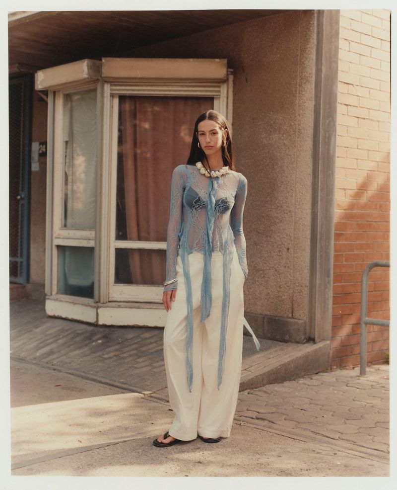 A full shot captures a young woman standing on a sidewalk in front of a brick building, wearing a sheer top and wide-leg pants.