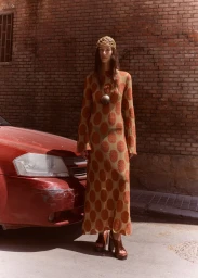 Model standing outdoors next to a red car wearing a patterned long sleeve dress and net headpiece