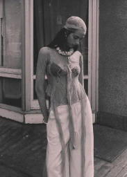 Model wearing a sheer lace long-sleeve top, matching headscarf, and wide-leg trousers standing outside in black and white.