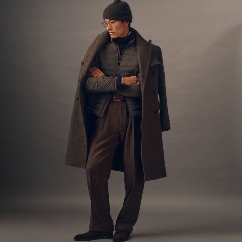 Man wearing a brown wool overcoat, puffer vest, and beanie standing with arms crossed against a plain grey studio background.
