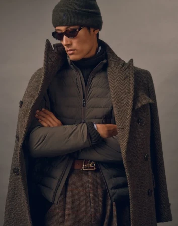 East Asian male model wearing dark sunglasses, an olive beanie, a quilted jacket, and a herringbone overcoat with arms crossed.