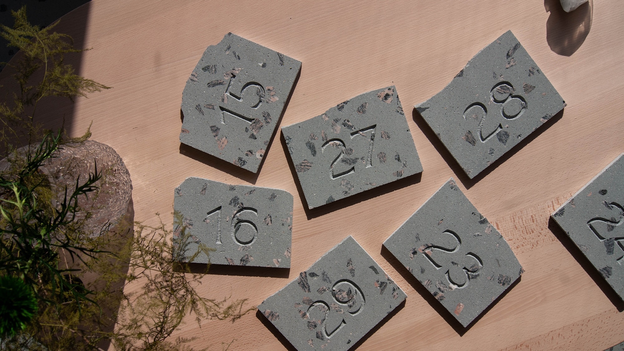 Engraved grey terrazzo stone number tiles arranged on a light wooden surface with botanical accents and natural shadows.