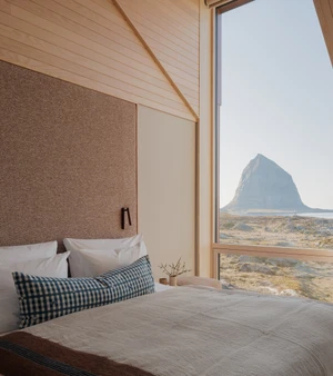A neatly made bed beside a tall window framing a solitary mountain peak and coastal landscape in a minimalist wood-paneled room.