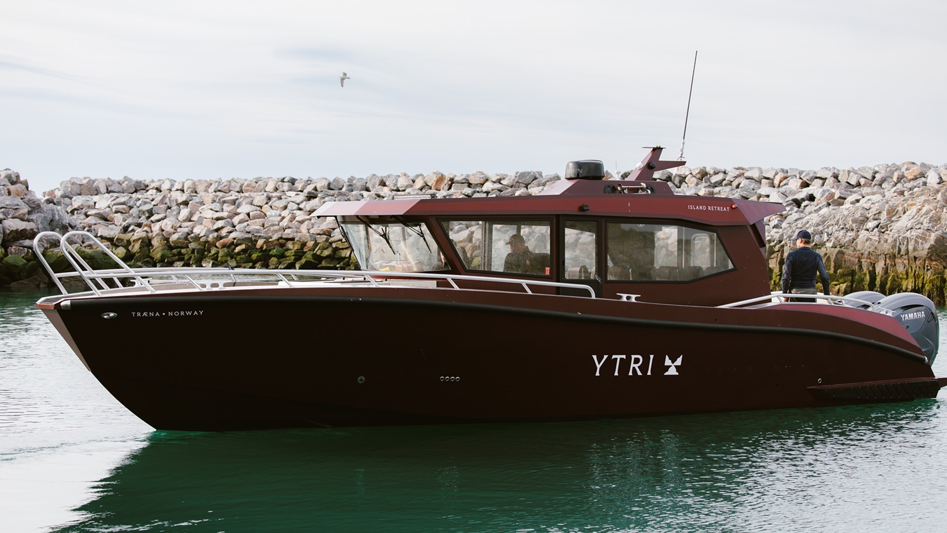 Dark burgundy Ytri boat floating on calm coastal water beside a rocky stone breakwater under overcast skies.