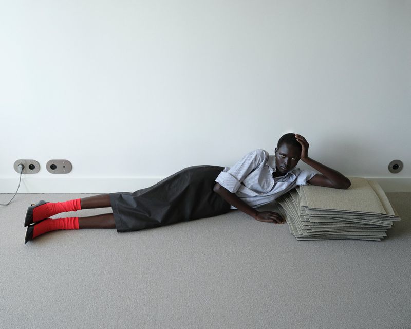 A person with dark skin lying on a grey carpet floor, wearing a white button-down shirt, a dark skirt, and vibrant red socks.