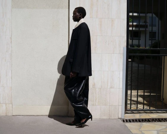 Model Aluel Keror in profile wearing an oversized black blazer and glossy wide-leg black trousers against a pale tiled wall.