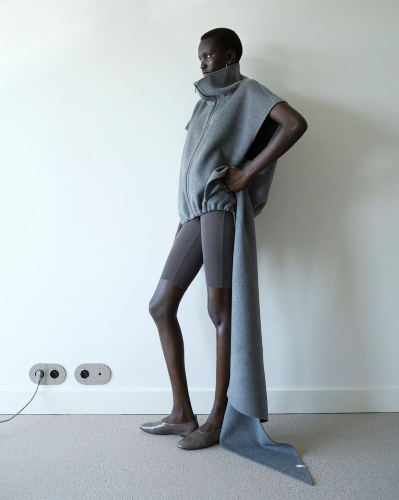Model Aluel Keror wearing an oversized grey zip-up sleeveless fleece with a trailing draped scarf panel and grey cycling shorts.