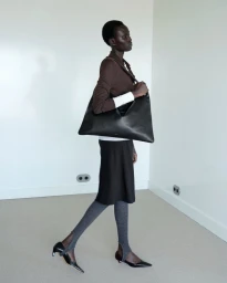 Model Aluel Keror walks in profile carrying an oversized black leather shoulder bag against a minimal white wall.