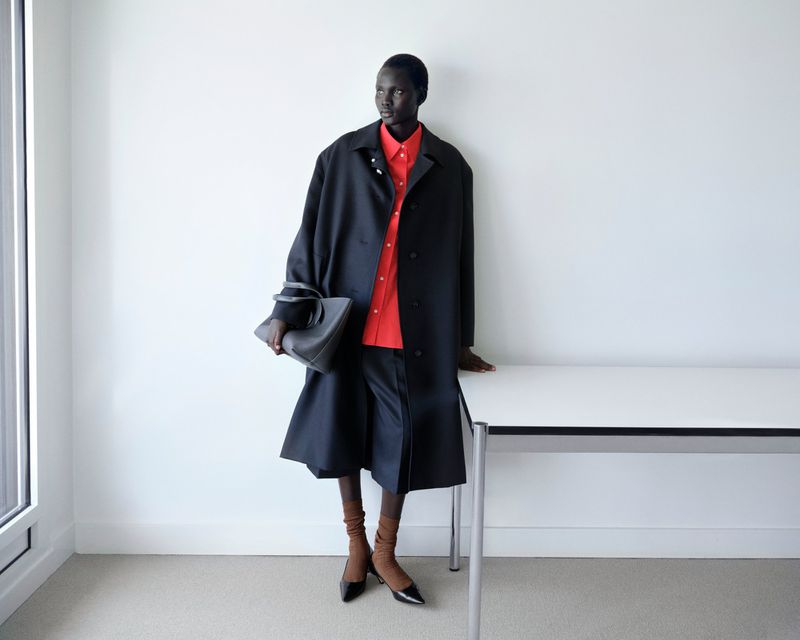 Aluel Keror wearing an oversized black coat, red shirt, and pointed-toe heels holding a leather bag beside a white desk.