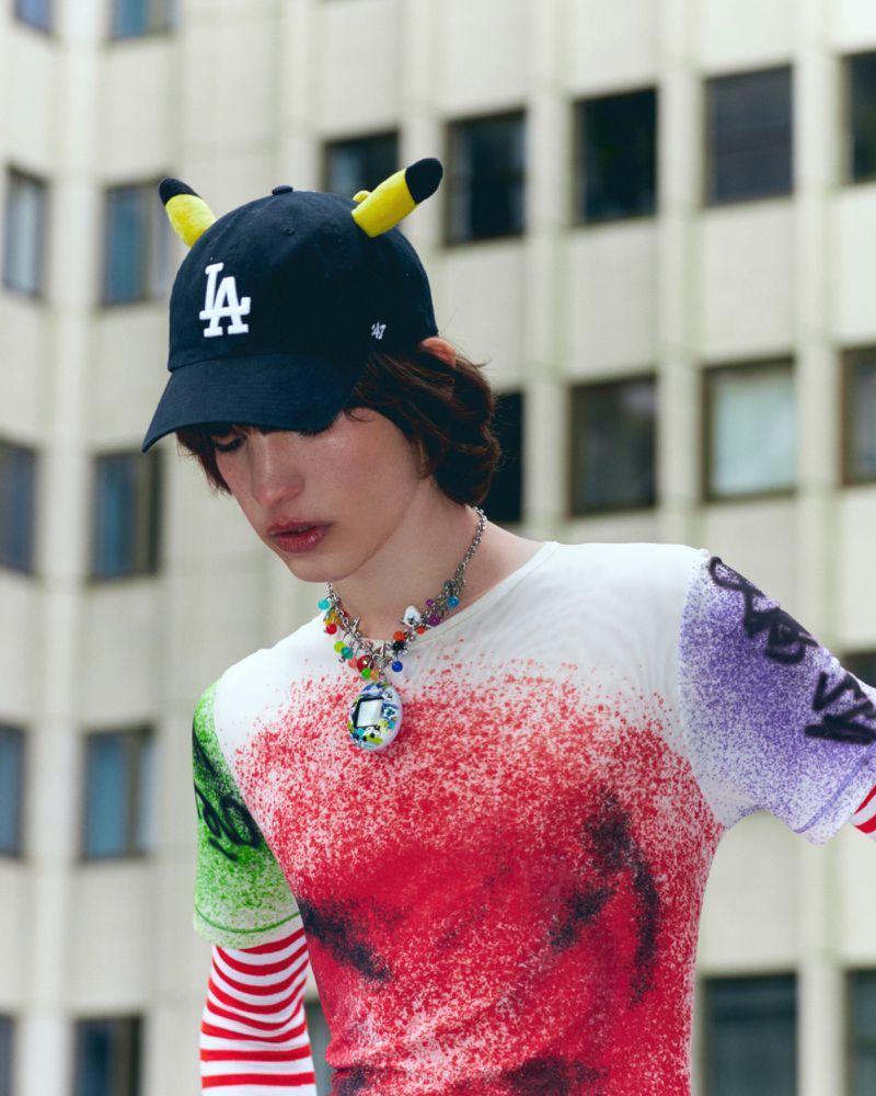 Young adult in graphic t-shirt, baseball cap with Pikachu ears, and beaded necklace standing in front of building exterior.