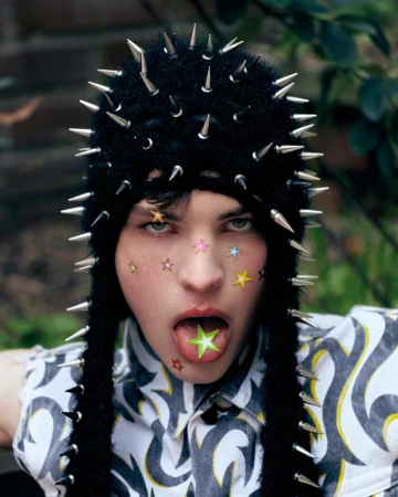 Portrait of a young adult with colorful star stickers on their face, wearing a black studded hat and printed shirt outdoors.
