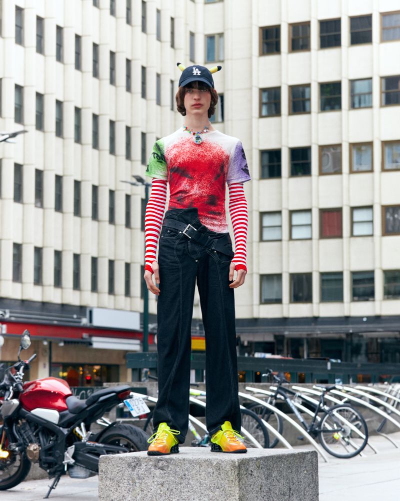 Standing in an urban setting, a young adult wears a baseball cap, patterned long-sleeve tee, dark jeans, and brightly colored sneakers.