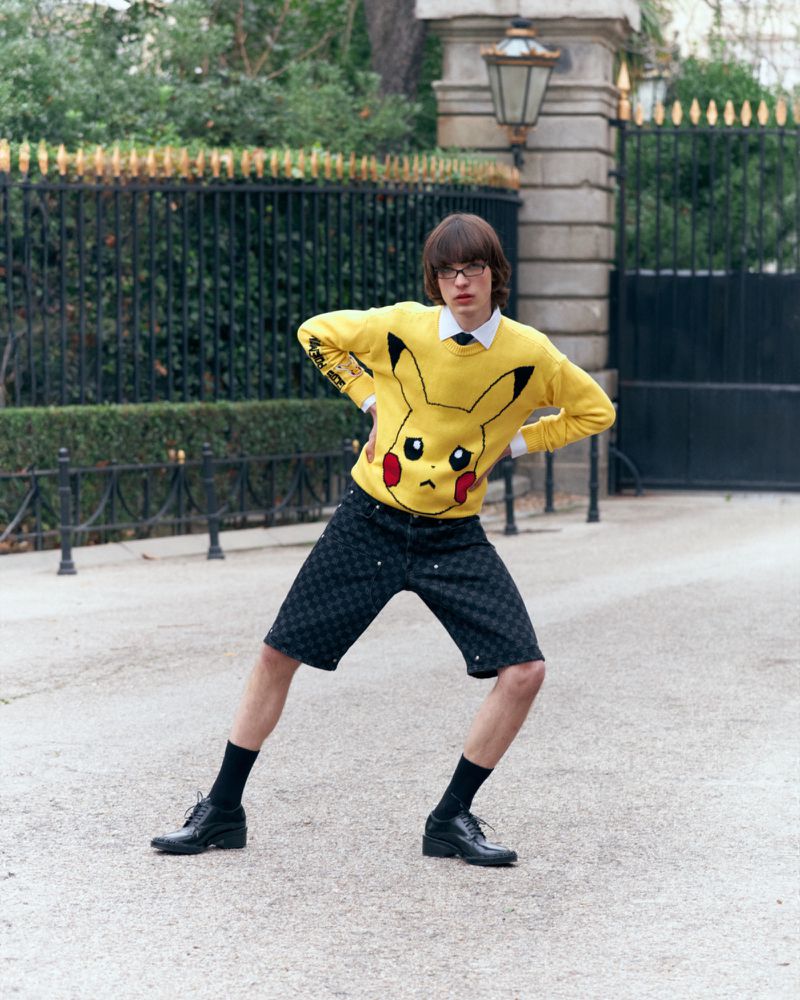 Fashion model poses outdoors in a yellow Pikachu sweater, black shorts, socks, and shoes in an urban setting near a gated stone building.