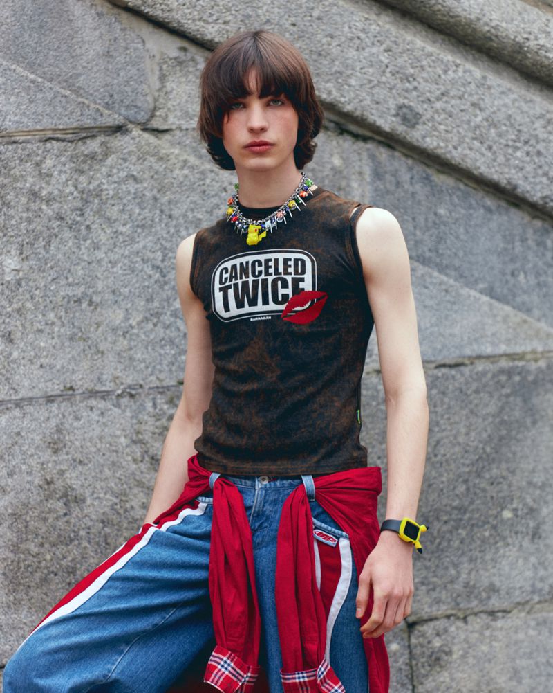 A young adult male model poses in a sleeveless graphic tee, jeans, and a colorful necklace against a stone background, fashionably styled.
