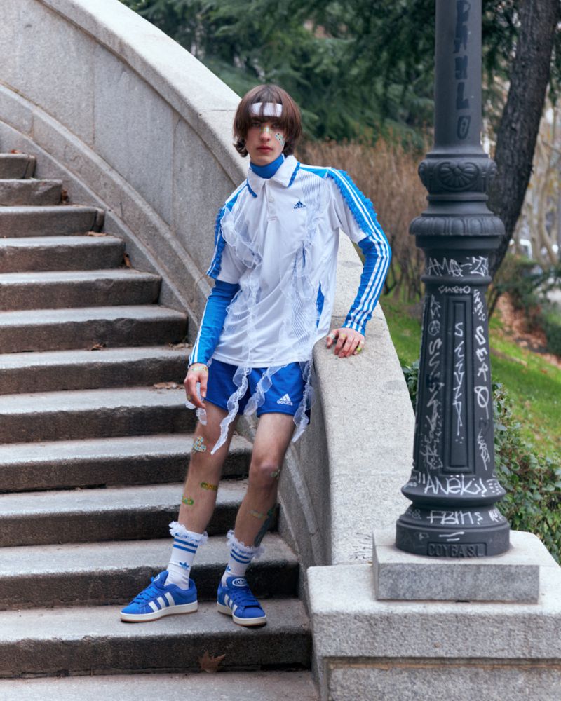 Young adult male model in Adidas sportswear poses on stone stairs outdoors, featuring a white and blue athletic outfit.