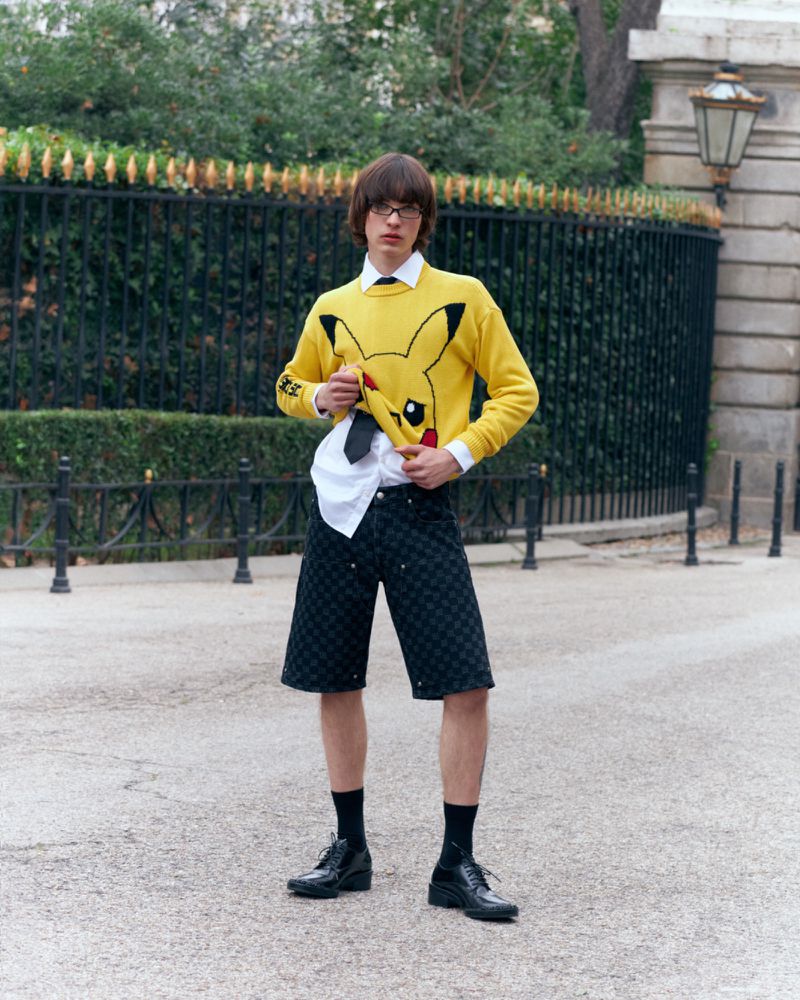A young man stands outside, wearing a yellow Pikachu sweater, black shorts, and dress shoes against a wrought-iron fence.