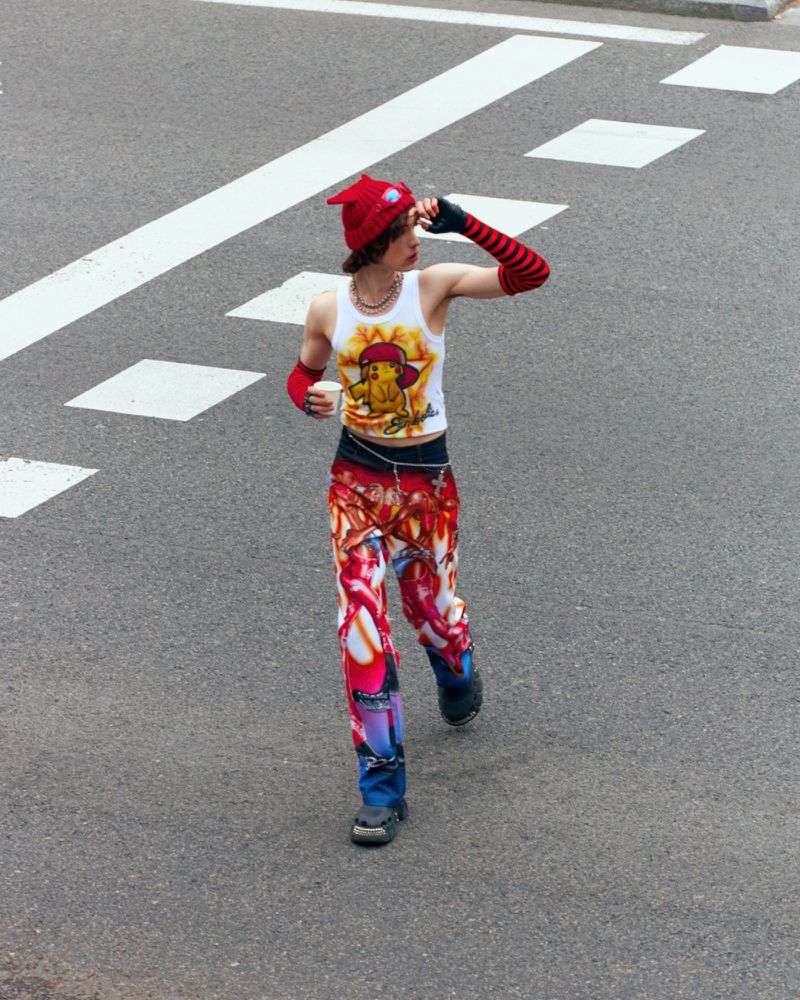 Young adult in knit hat and patterned clothes walks in a road with white markings, holding a cup, viewed from a high angle.