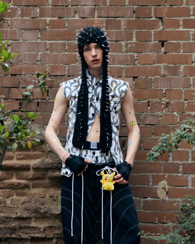 Adult male poses against a brick wall wearing a spiked hat and vest with a Pokemon doll accessory and black pants with white stripes.