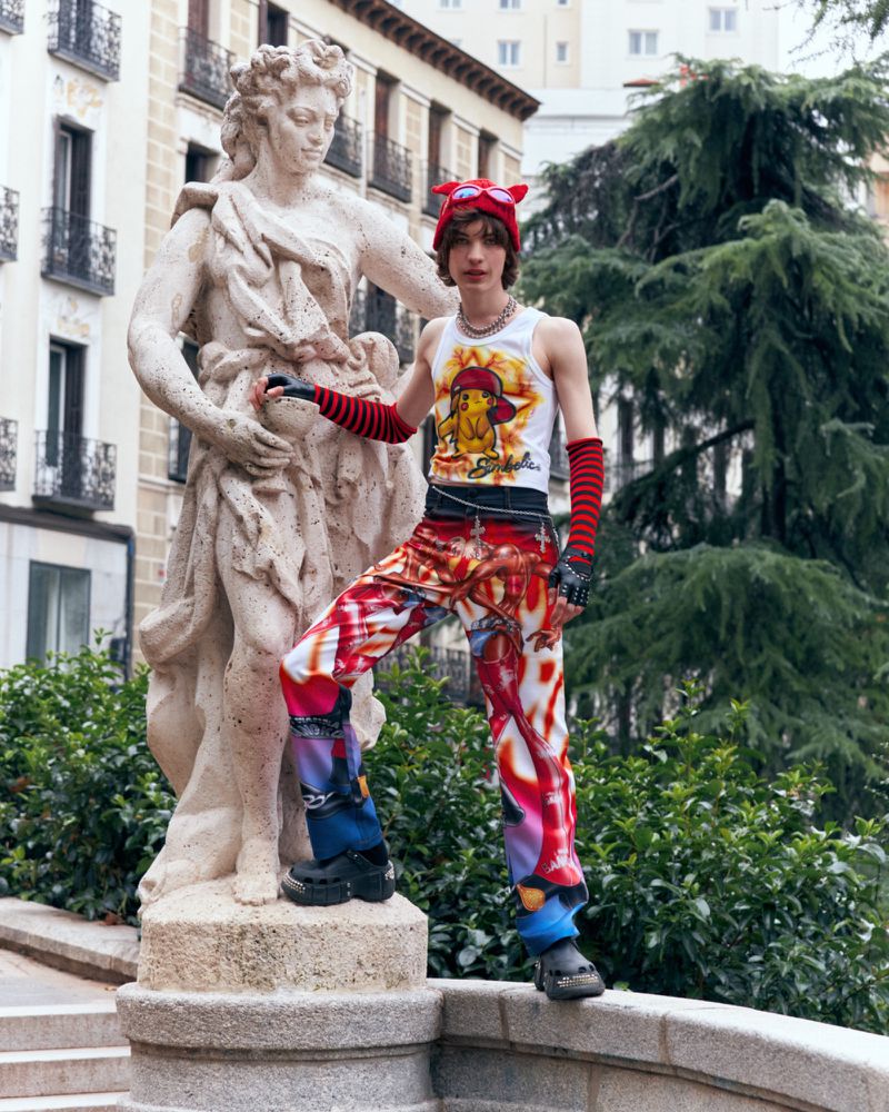 A young adult stands in front of a statue in a park-like setting, wearing a hat, graphic tank top, and colorful patterned pants.