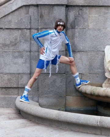 A person in an Adidas outfit poses on a fountain with gray stone walls in the background, creating a unique fashion statement.