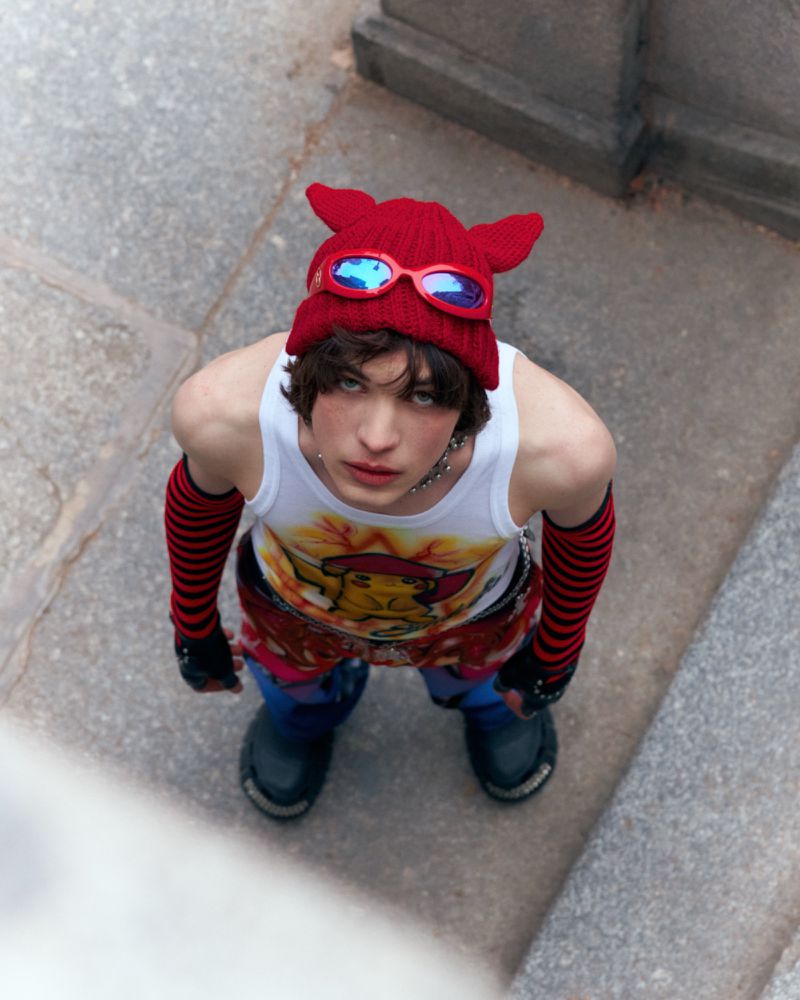 A young adult in red knit cap and graphic tank top looks up at the camera outdoors with a light-grey stone background.