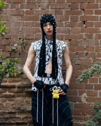 Young man wearing a black spiked beanie, patterned vest, and dark pants standing against a weathered brick wall.