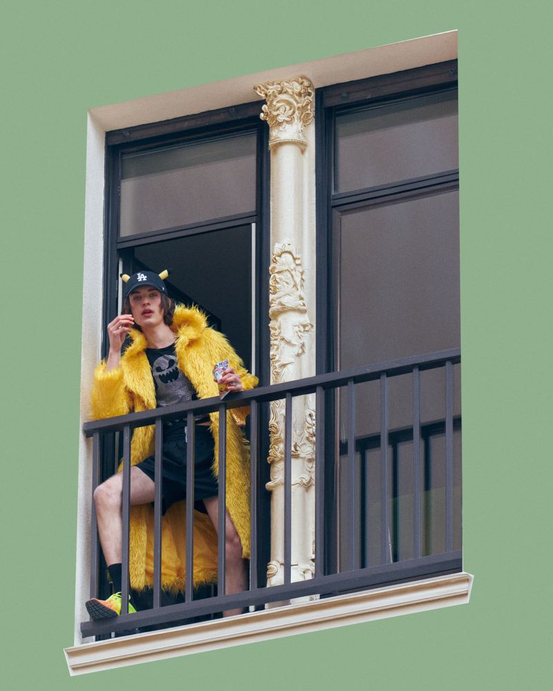 A young man wearing a yellow faux fur coat and horned baseball cap standing on a balcony with ornate columns.