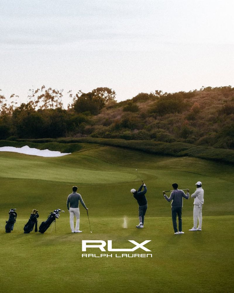 Four male golfers in athletic wear standing on a golf fairway with one mid-swing and golf bags nearby.
