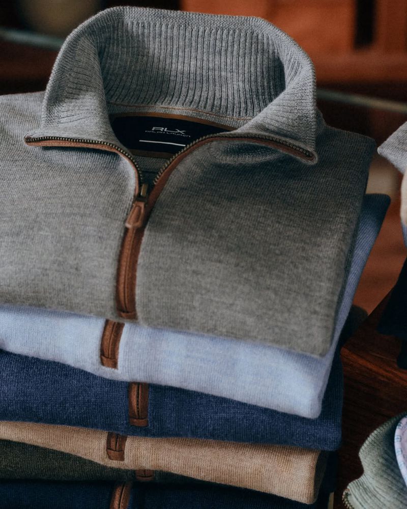 A stack of folded RLX Ralph Lauren quarter-zip sweaters in assorted colors with brown zipper detailing.