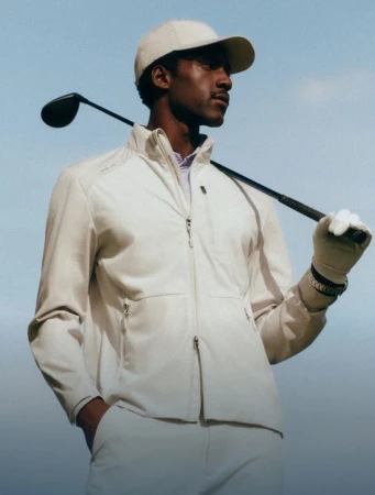 Black male model in a beige zip jacket and cap resting a golf club over his shoulder against a blue sky.