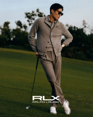 Man in a grey knit cardigan and pleated trousers holding a golf club on a green course for Ralph Lauren RLX.