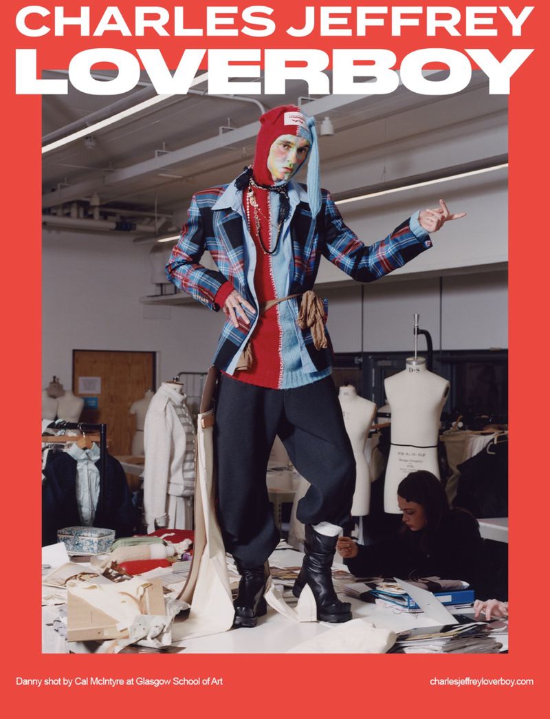 Model in Charles Jeffrey Loverboy plaid jacket and knit bonnet poses in a sewing studio with dress forms and design materials.